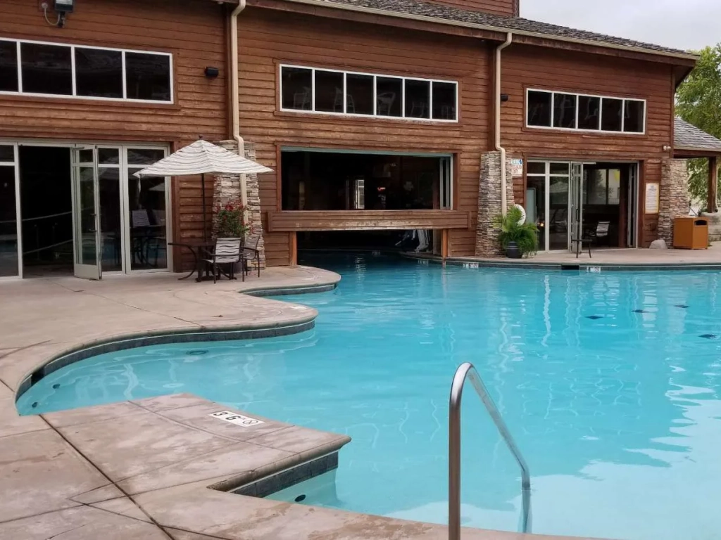 Outdoor resort pool with turquoise water beside a rustic log and stone lodge building with patio seating.