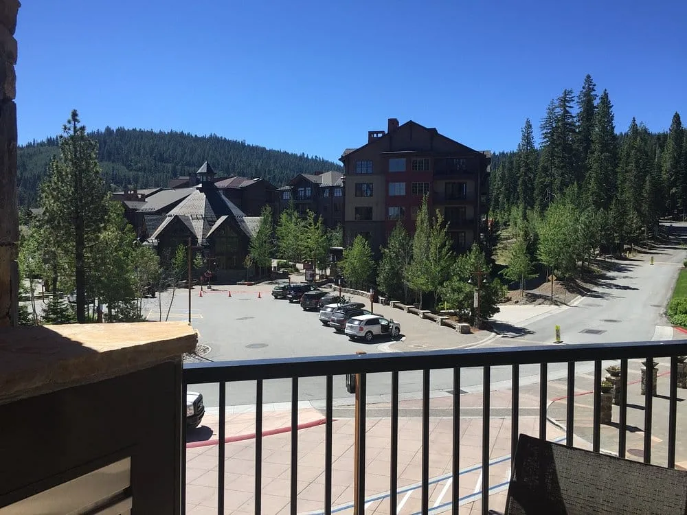 Balcony view overlooking a sunny mountain resort parking area with pine trees and lodge-style buildings.