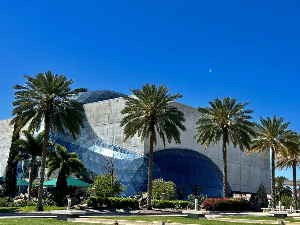 The Salvador Dalí Museum in St. Petersburg Florida with its dramatic curved glass façade surrounded by tall palm trees under a clear blue sky.
